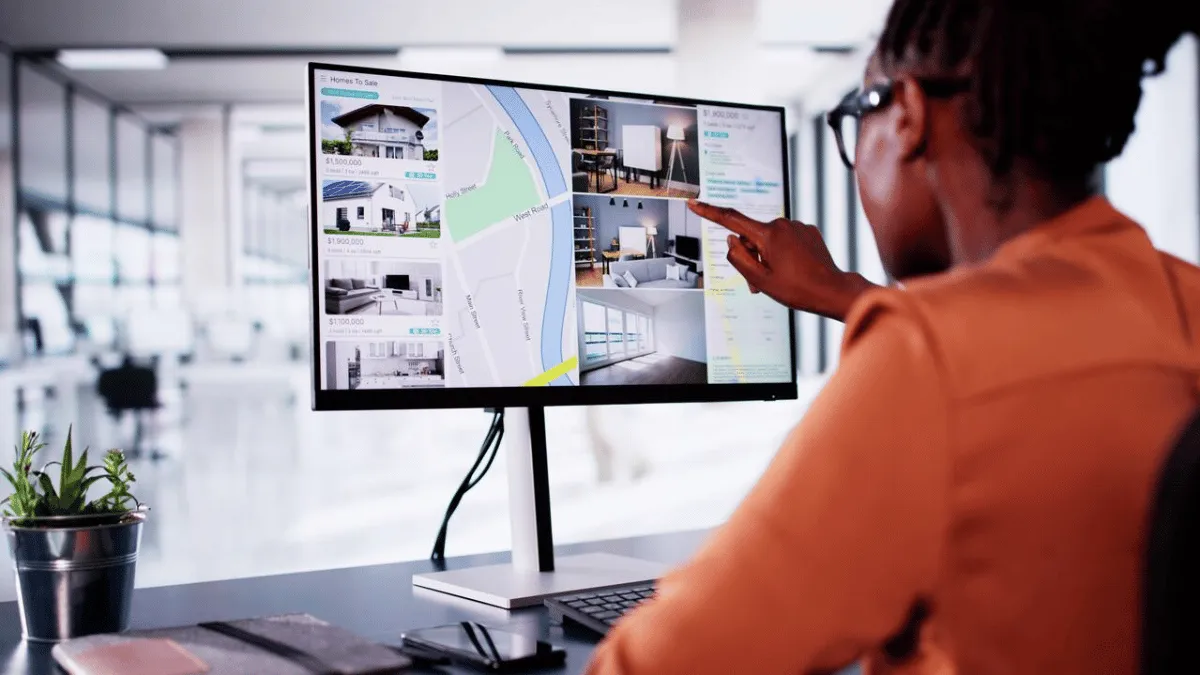 A person with dark skin reviews real estate listings on a computer in a bright office setting, pointing at an interior photo of a house for sale.