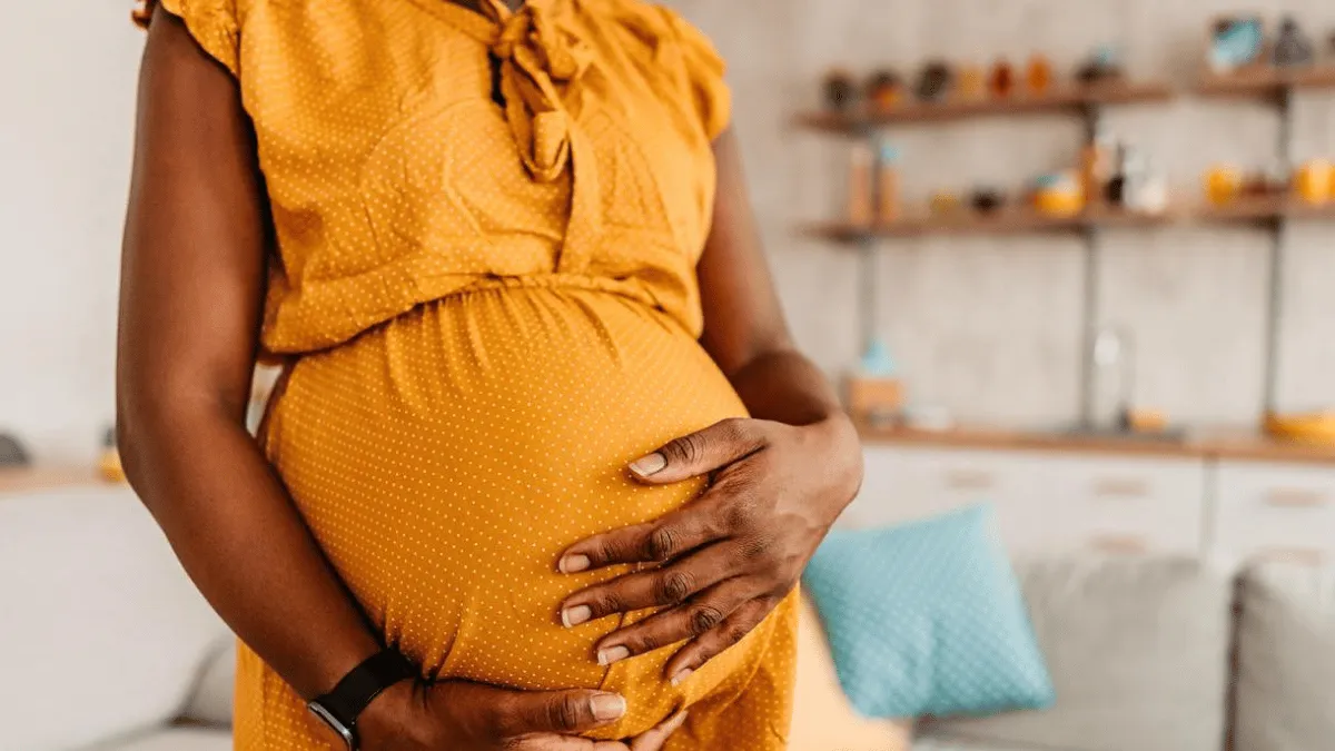 A person with dark skin, wearing a mustard-yellow dress, gently holds their pregnant belly with both hands.