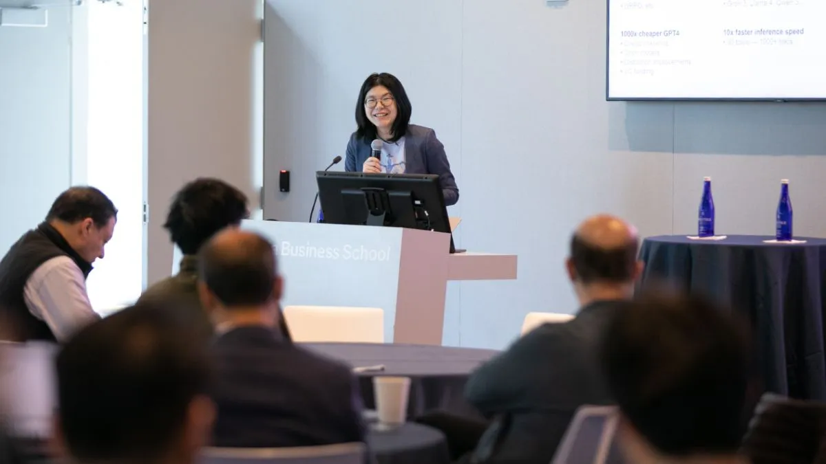 Presenter standing at a podium labeled “Business School,” speaking to an audience in a conference room, with presentation slides on a screen in the background.