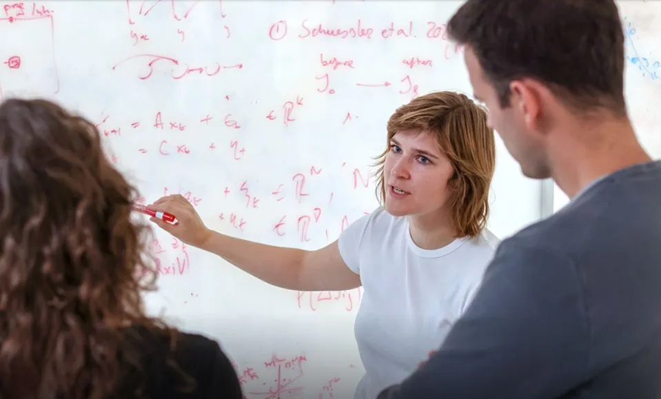 Lea Duncker at whiteboard explaining equations to two colleagues 