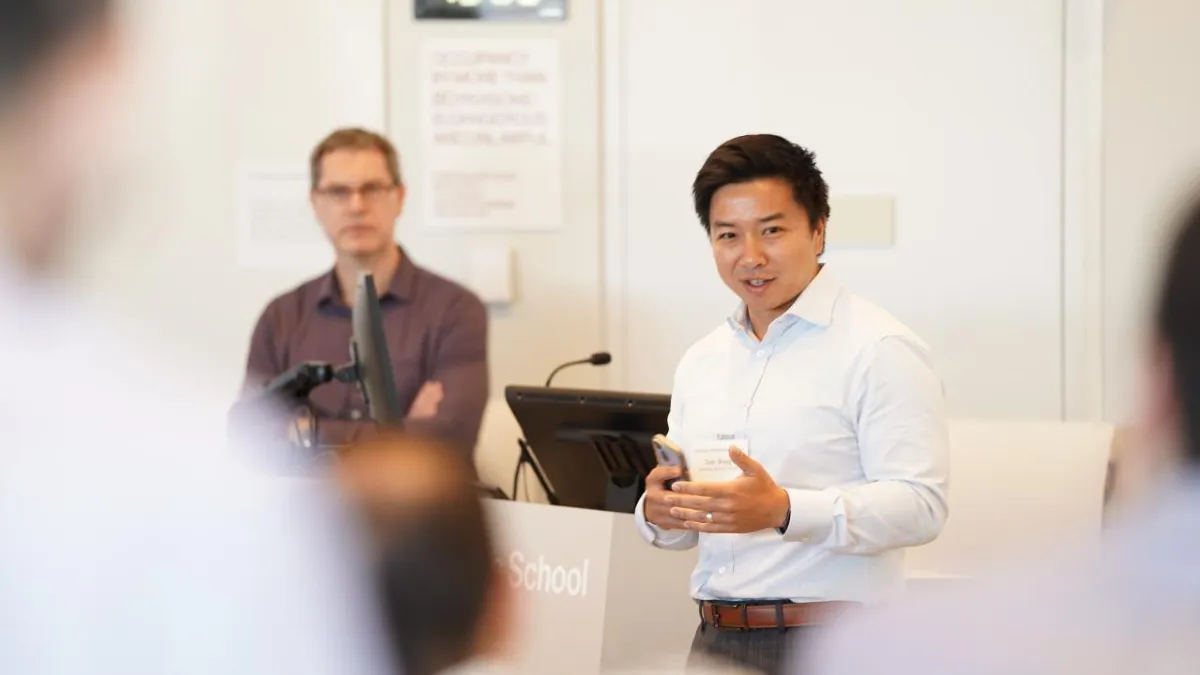 A speaker in a white shirt addresses an audience during a classroom discussion, gesturing while speaking. Another person stands in the background near a podium and computer setup, listening attentively. The setting appears to be an academic lecture or workshop.