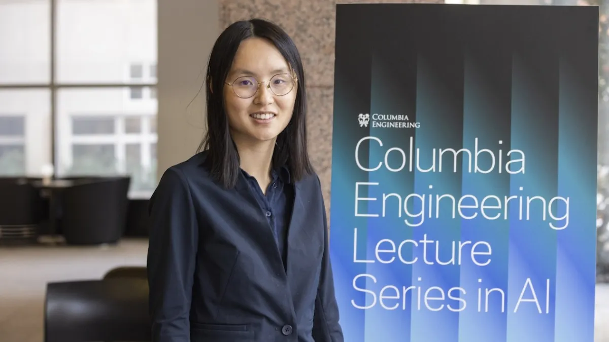 A woman in a dark blazer stands next to a sign for the “Columbia Engineering Lecture Series in AI,” smiling at the camera in a modern indoor setting.