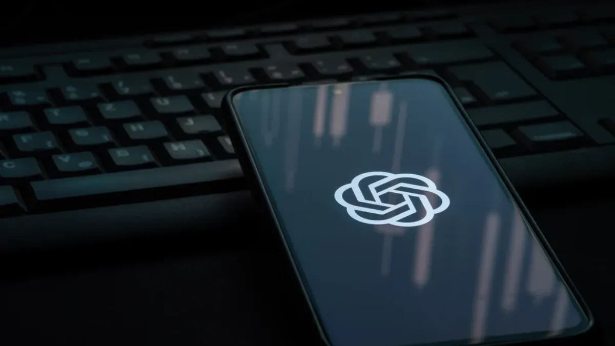 A phone is on a laptop keyboard, the phone screen is black with only the ChatGPT logo visible in the middle of the screen. 