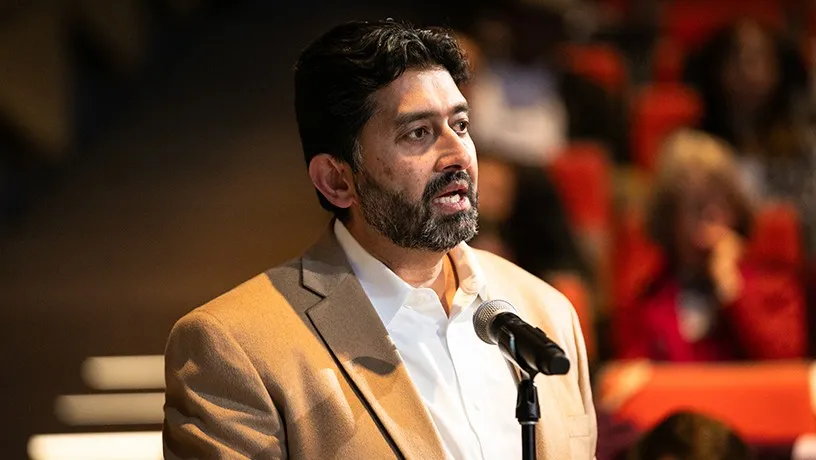 A male audience member speaking into a microphone. Other audience members sit in the background out of focus.