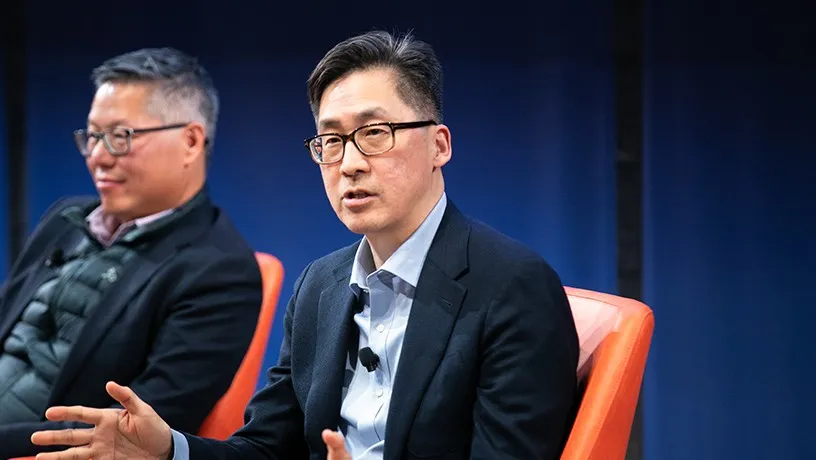 An Asian panelist addressing the crowd from his seat. Another male co-panelist sits in the background
