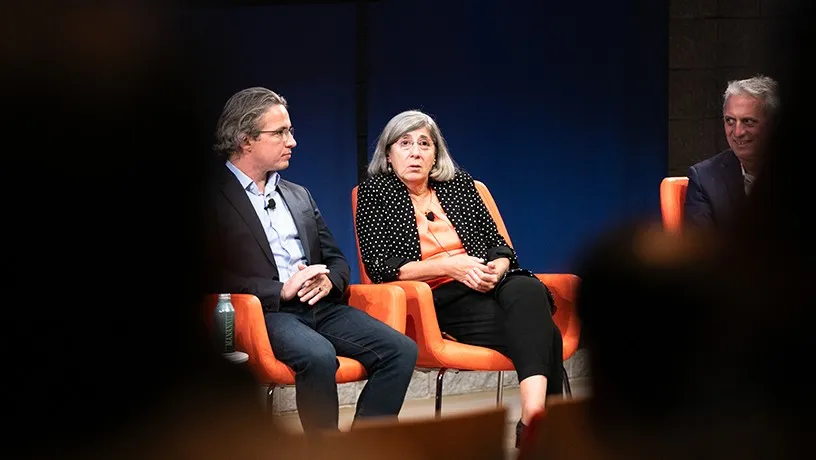 Three panelists, two males and a female, sitting on chairs on stage. The backs of audience members' heads in the foreground obstruct a view of the stage.