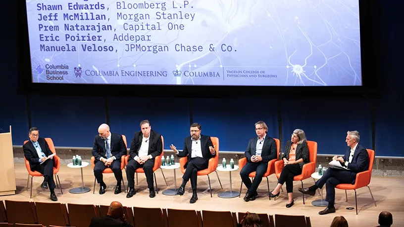 Seven panelists, six males and one female, sit in on-stage chairs. A screen behind the panelists reads names and their affiliations: Shawn Edwards, Bloomberg L.P., Jeff McMillan, Morgan Stanley; Prem Natarajan, Capital One; Eric Poirier, Addepar; Manuela Veloso, JPMorgan Chase & Co. The central male panelists gestures to the audience sitting in front of the stage. 