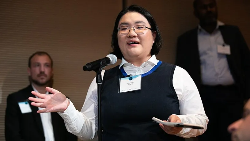 A female audience member gesturing with their palms open and speaking into a microphone. Other audience members stand in the background