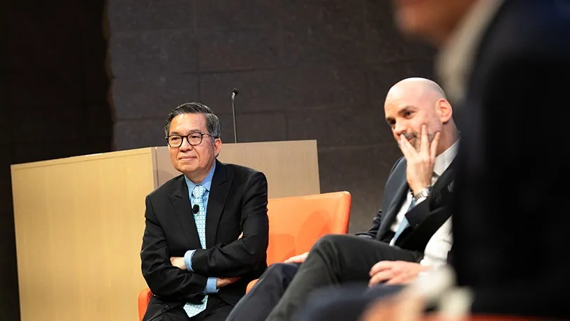 Shih-Fu Chang and Shawn Edwards sitting in chairs on-stage. A person in the foreground partially blocks a view of the stage.